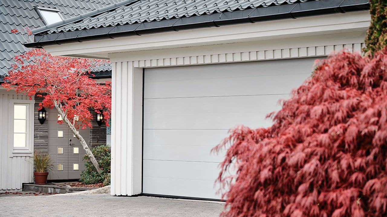 Lys garasjeport på hvit garasje med trær med rosa blader rundt.
