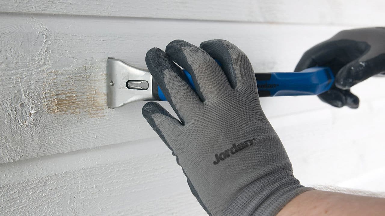 Nærbilde av hender med arbeidshansker som skraper av maling på fasaden før maling utvendig.